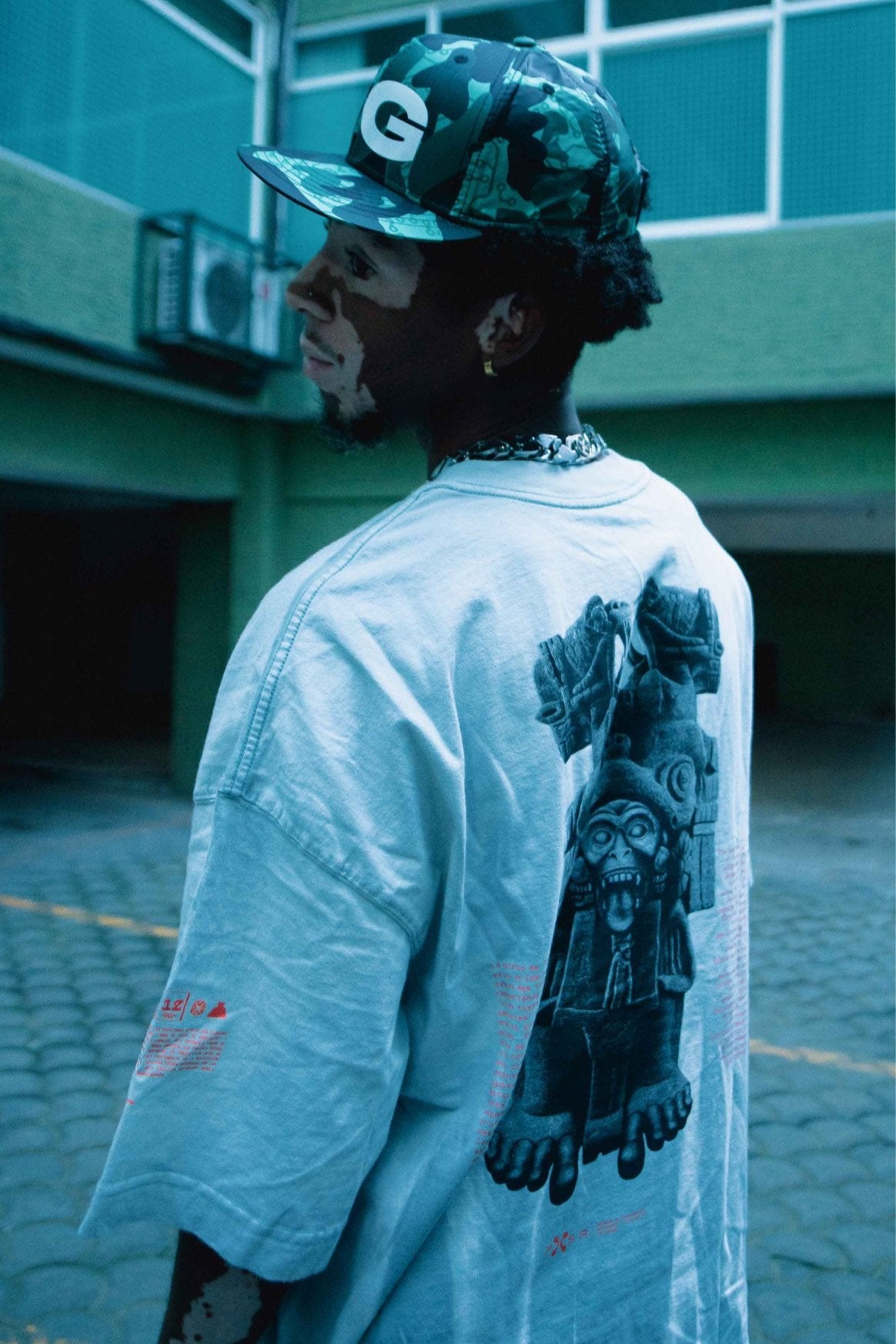 A person with a distinctive skin pattern, evocative of the Coatlicue design by Machina - Wearable Technology, dons a cap emblazoned with a G logo and a relaxed gray shirt showcasing a graphic design on the back. They are positioned in an outdoor scene featuring green walls and large windows in the background.