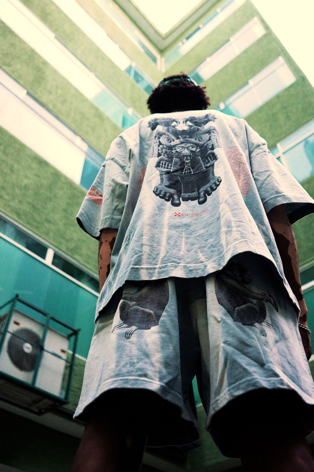 An individual in loose-fitting apparel embellished with cartoon motifs stands before a green structure, evoking the majestic presence of the Coatlicue. The upward angle reveals the expansive windows of the modern design set against a cloudy sky, suggesting hidden twin realms above. (Coatlicue by Machina - Wearable Technology)