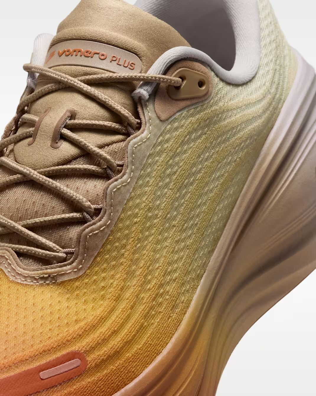 A close-up of the Nike Vomero Plus “Cheat Day” athletic shoe by Machina Wearable Technology in beige and orange, featuring VOMERO PLUS branding on the tongue, detailed stitching, laces, textured fabric, and a ZoomX midsole engineered for ultimate comfort in a running shoe.