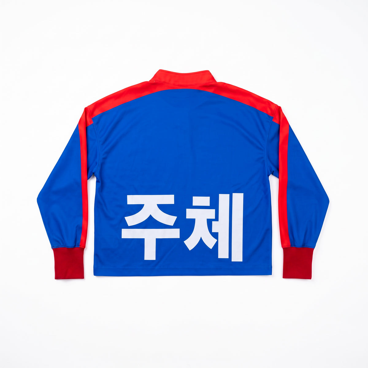 A long-sleeved blue jacket from Machina - Wearable Technology, called COREA DEL NORTE, featuring red detailing on the collar, shoulders, and cuffs and displayed flat. The back showcases large white Korean characters in a bold, propaganda-inspired football shirt style.