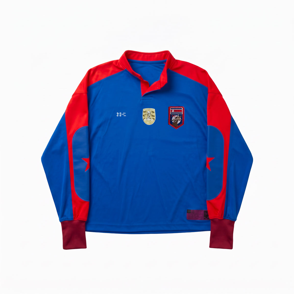 The COREA DEL NORTE jersey by Machina - Wearable Technology is a long-sleeved blue soccer shirt featuring red shoulders, maroon cuffs, and two embroidered crests on the chest. This concept piece is inspired by classic North Korean football jerseys and is shown laid flat on a white background.