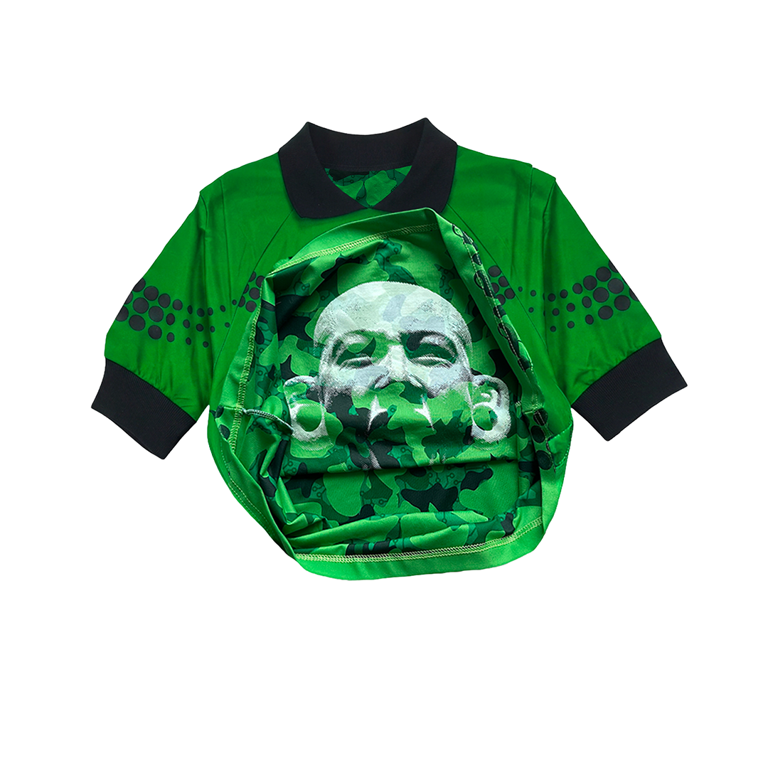 The MEXICANOS AL GRITO DE GUERRA by Machina - Wearable Technology is a green collared shirt with black trim and dotted sleeves, folded open to reveal a hidden stone face image camouflaged in green patterns.