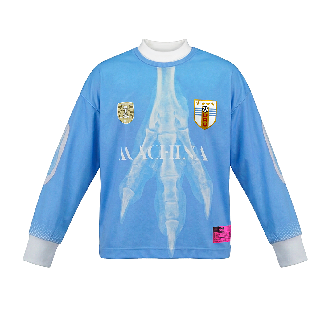 The LA GARRA jersey by Machina - Wearable Technology is a blue long-sleeve top with white cuffs, a bird claw x-ray image on the front, two chest badges, MACHINA in white, and a pink hem label—evoking classic World Cup goalkeeper jerseys.