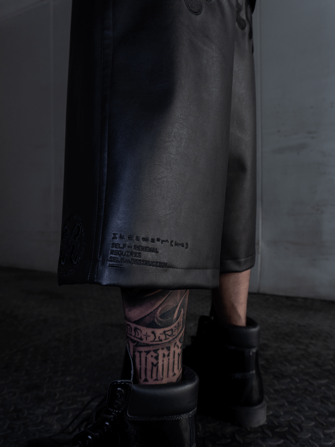 A person models the Machina - Wearable Technology All-Black Eco Leather Nagamani Set with black boots, standing on a textured metal floor. Part of their tattooed leg is visible. The background is plain and industrial-looking.