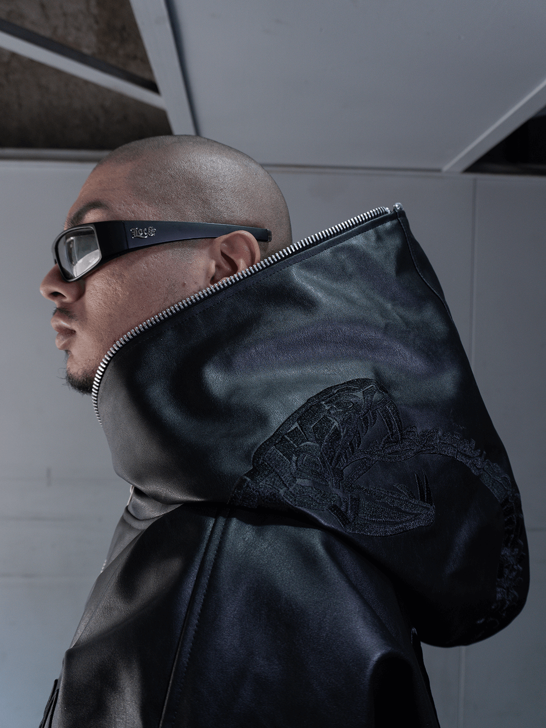 A person with a shaved head wears thick black sunglasses and the Machina - Wearable Technology All-Black Eco Leather Osseous Full Zipper set, standing indoors against a geometric ceiling.