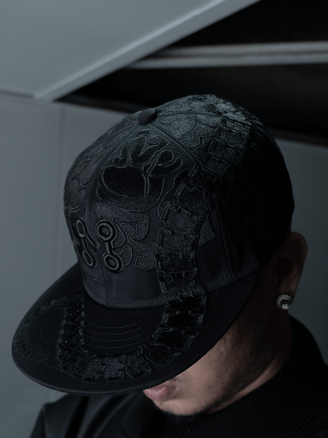 A person dressed in the All-Black Eco Leather Nagamani Set by Machina - Wearable Technology, wearing a textured black cap low over their face, an earring visible, stands against a muted background in dark attire.
