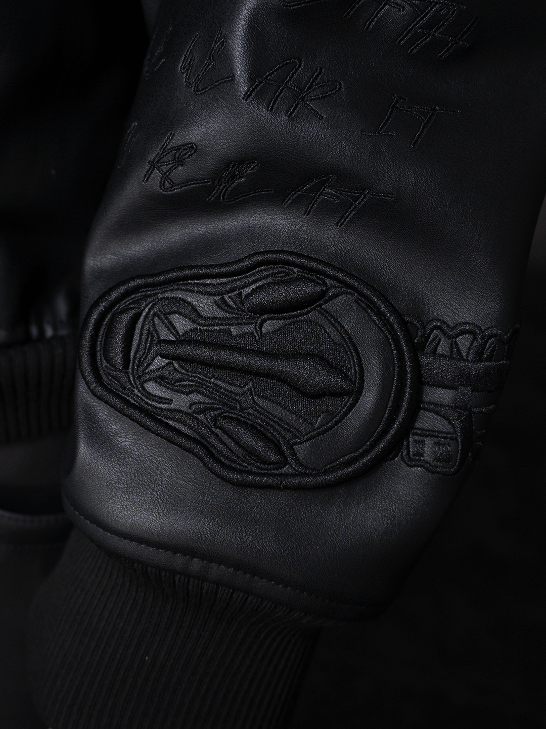 Close-up of the All-Black Eco Leather Nagamani Set by Machina - Wearable Technology: a black sleeve with intricate abstract embroidery, raised text details, and a matching ribbed cuff.