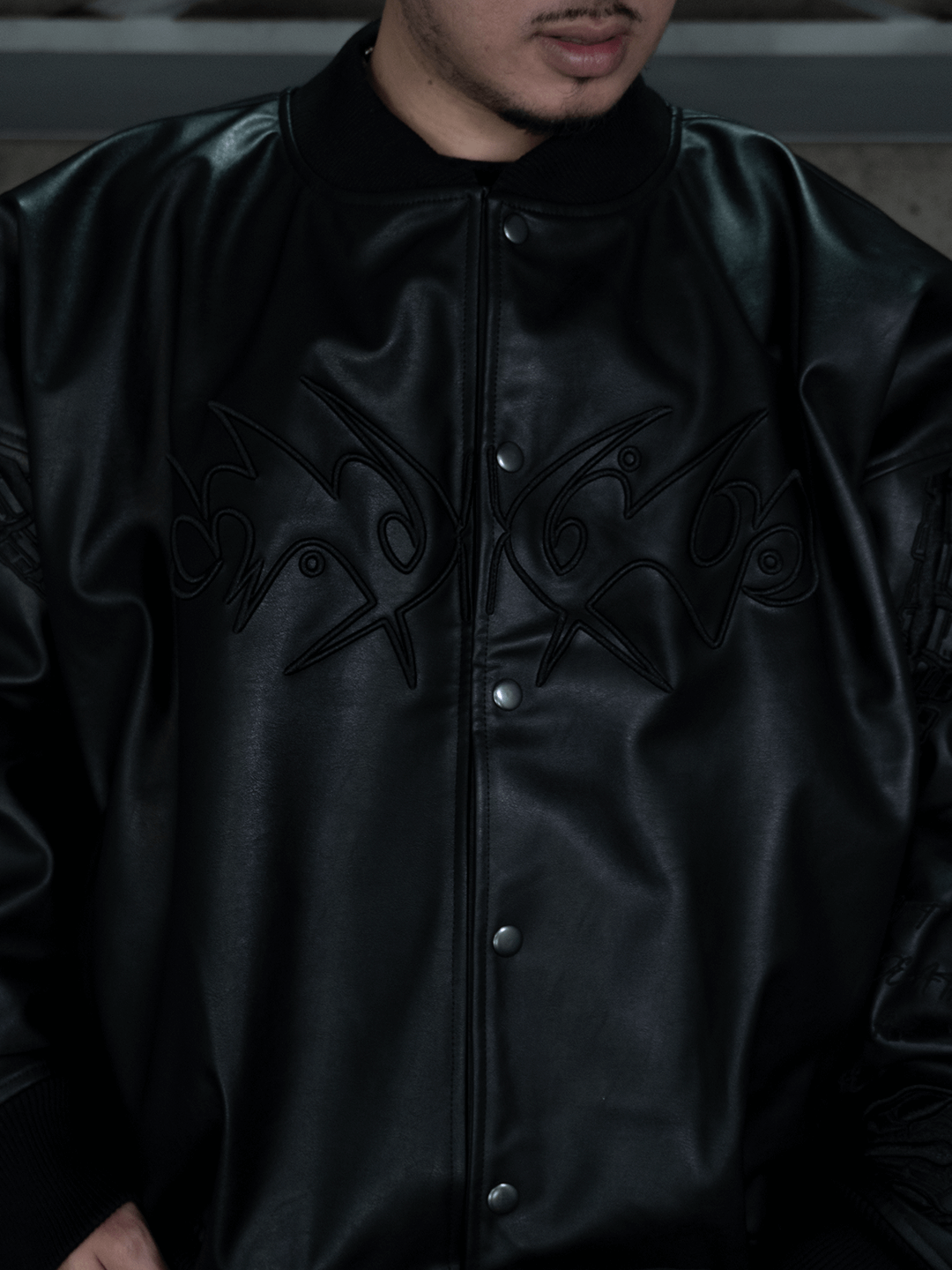 A person wearing the All-Black Eco Leather Nagamani Set by Machina – Wearable Technology, featuring abstract embroidery on the chest, stands indoors with only the lower half of their face visible.