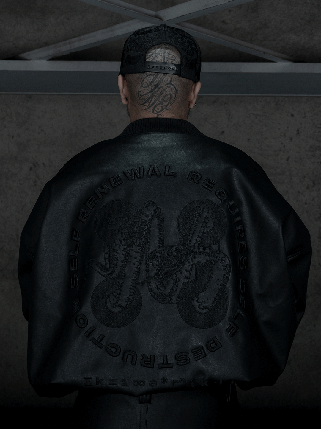A person with a tattooed head and black cap faces away, wearing the Machina - Wearable Technology All-Black Eco Leather Nagamani Set with bold SELF RENEWAL REQUIRES SELF DESTRUCTION embroidery and decorative detailing on the back.