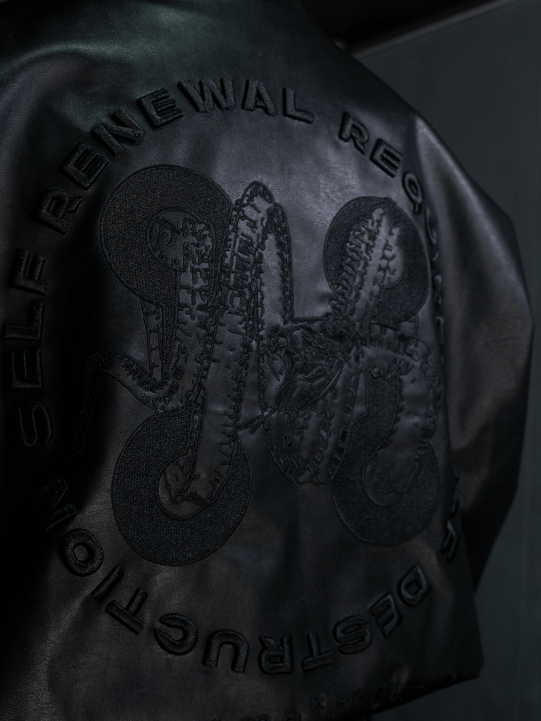 A close-up of the Machina - Wearable Technology All-Black Eco Leather Nagamani Set shows detailed black embroidery of abstract forms and the phrase "SELF RENEWAL REQUIRES DESTRUCTION" arranged in a circular pattern.