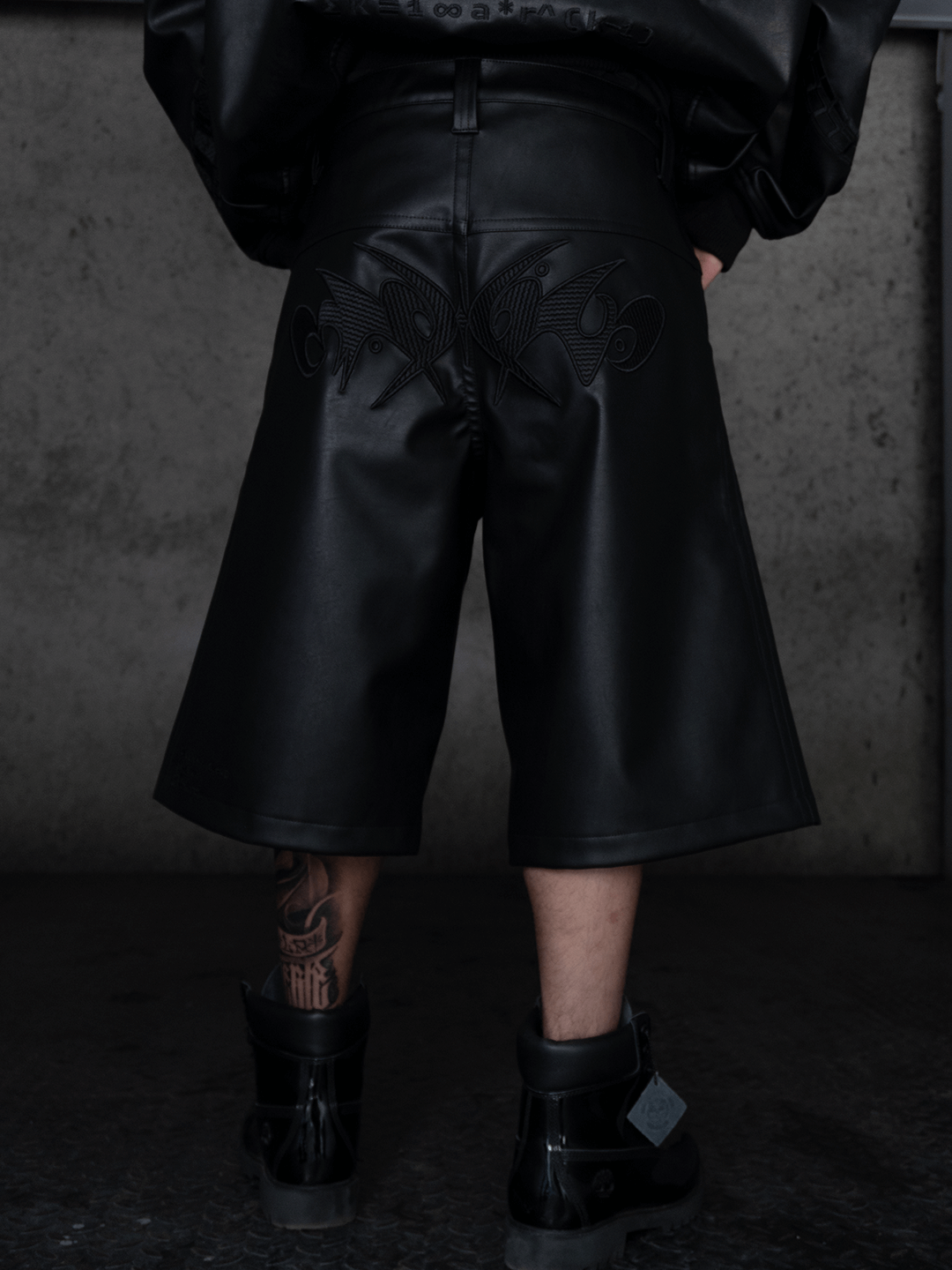 A person faces a concrete wall, wearing Machina - Wearable Technology's All-Black Eco Leather Nagamani Set with embroidered details, paired with black boots and displaying a tattoo on their left calf.