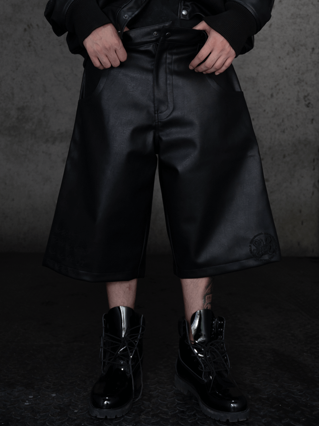 Wearing Machina – Wearable Technology's All-Black Eco Leather Nagamani Set, a person stands with hands in pockets against a textured gray wall, showing only their lower half clad in knee-length shorts and shiny lace-up boots.