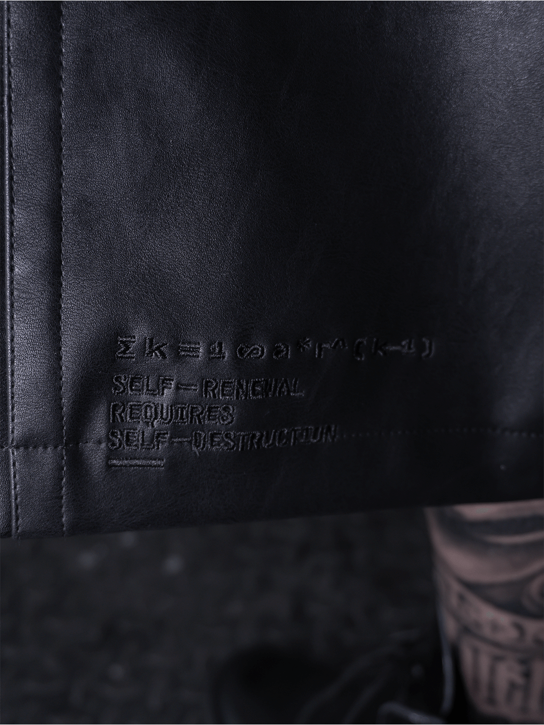 Close-up of the All-Black Eco Leather Nagamani Set by Machina - Wearable Technology, featuring embossed text: “SELF-RENEWAL REQUIRES SELF-DESTRUCTION,” with partially obscured text and symbols above.