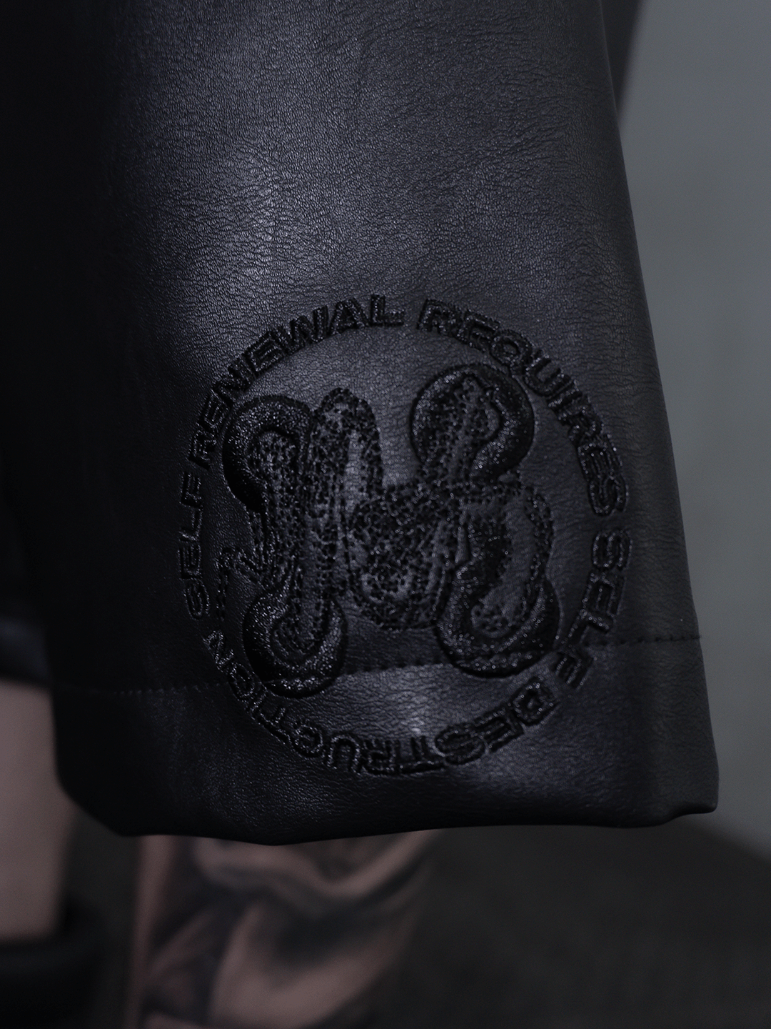A detail view of the All-Black Eco Leather Nagamani Set by Machina - Wearable Technology, showcasing an embroidered circular patch with “SELF RENEWAL REQUIRES SELF DESTRUCTION” around an abstract central logo.