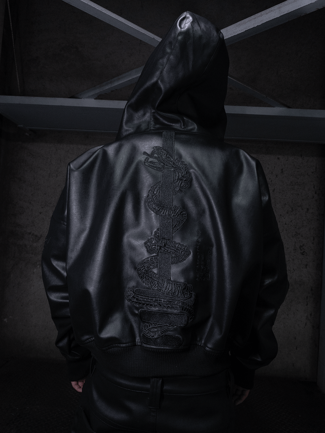 Wearing the All-Black Eco Leather Bubble Set by Machina - Wearable Technology, a person faces away in a dimly lit industrial space, showcasing an embossed snake design on the back of their hooded jacket.
