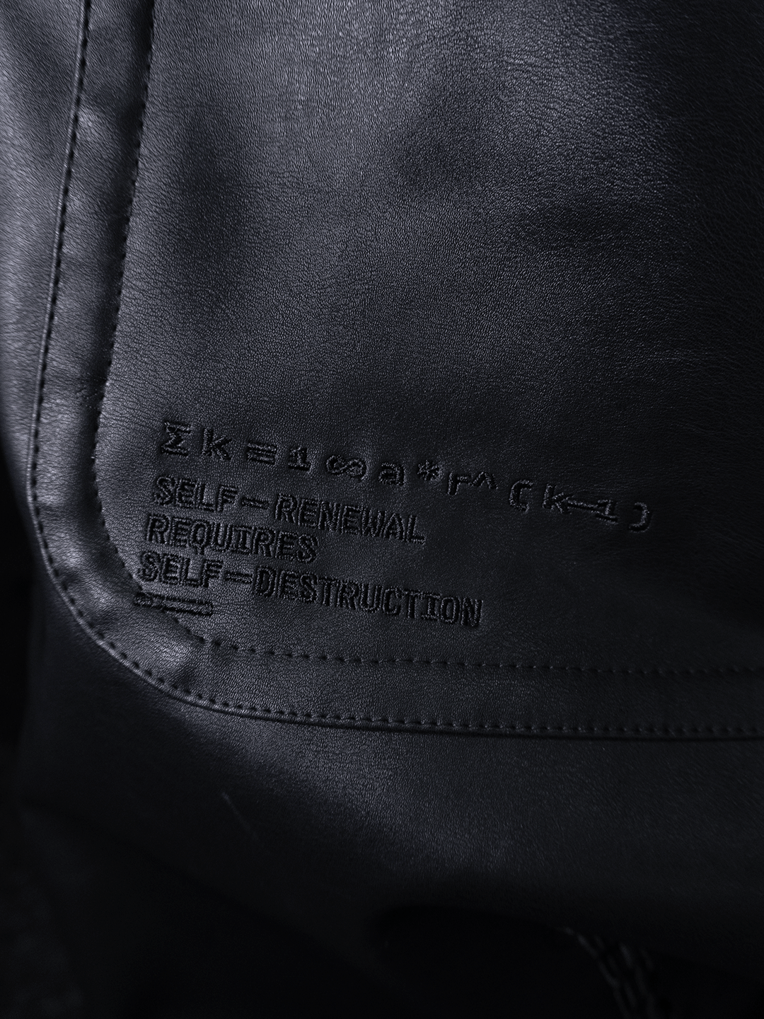 Close-up of All-Black Eco Leather Osseous Full Zipper set by Machina - Wearable Technology, featuring embroidered text "SELF-RENEWAL REQUIRES SELF-DESTRUCTION" and an equation above, with visible stitching and rich texture.