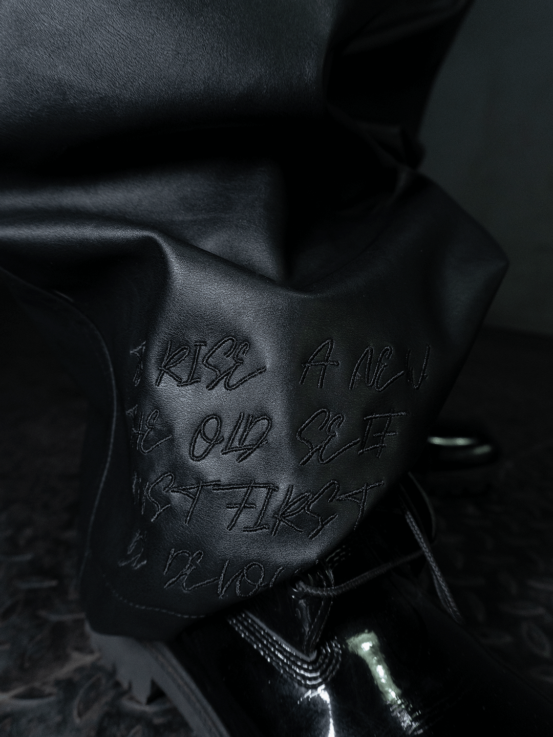 Close-up of the Machina - Wearable Technology All-Black Eco Leather Osseous Full Zipper set pants with embroidered "RISE A NEW AS OLD SELF FIRST," matched with black boots on a textured dark floor.