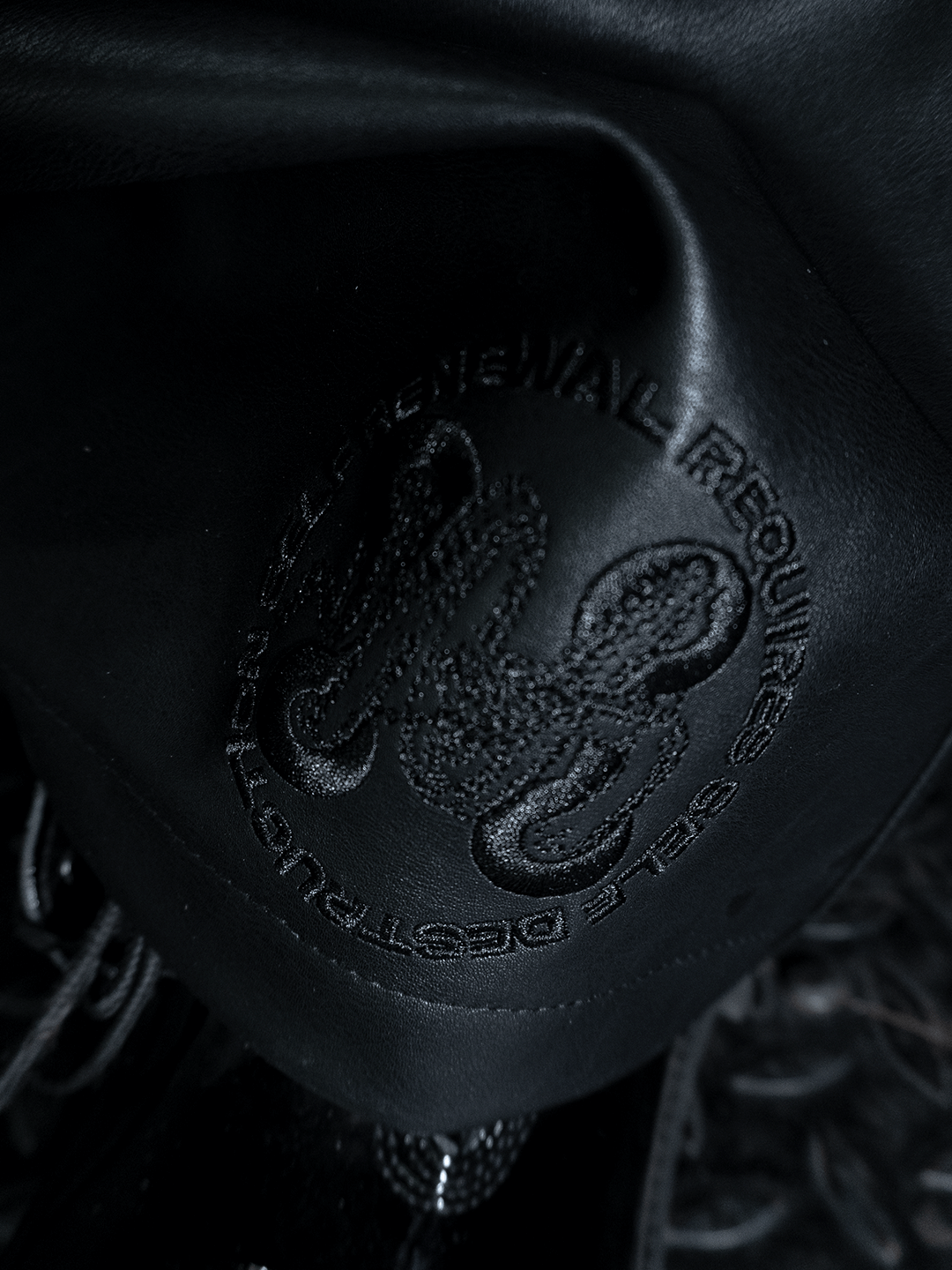 Close-up of the All-Black Eco Leather Osseous Full Zipper set by Machina - Wearable Technology, featuring embossed leather with a stylized serpent logo and the partially visible words “DESTROY” and “PROFESSIONAL REQUIRE.”.