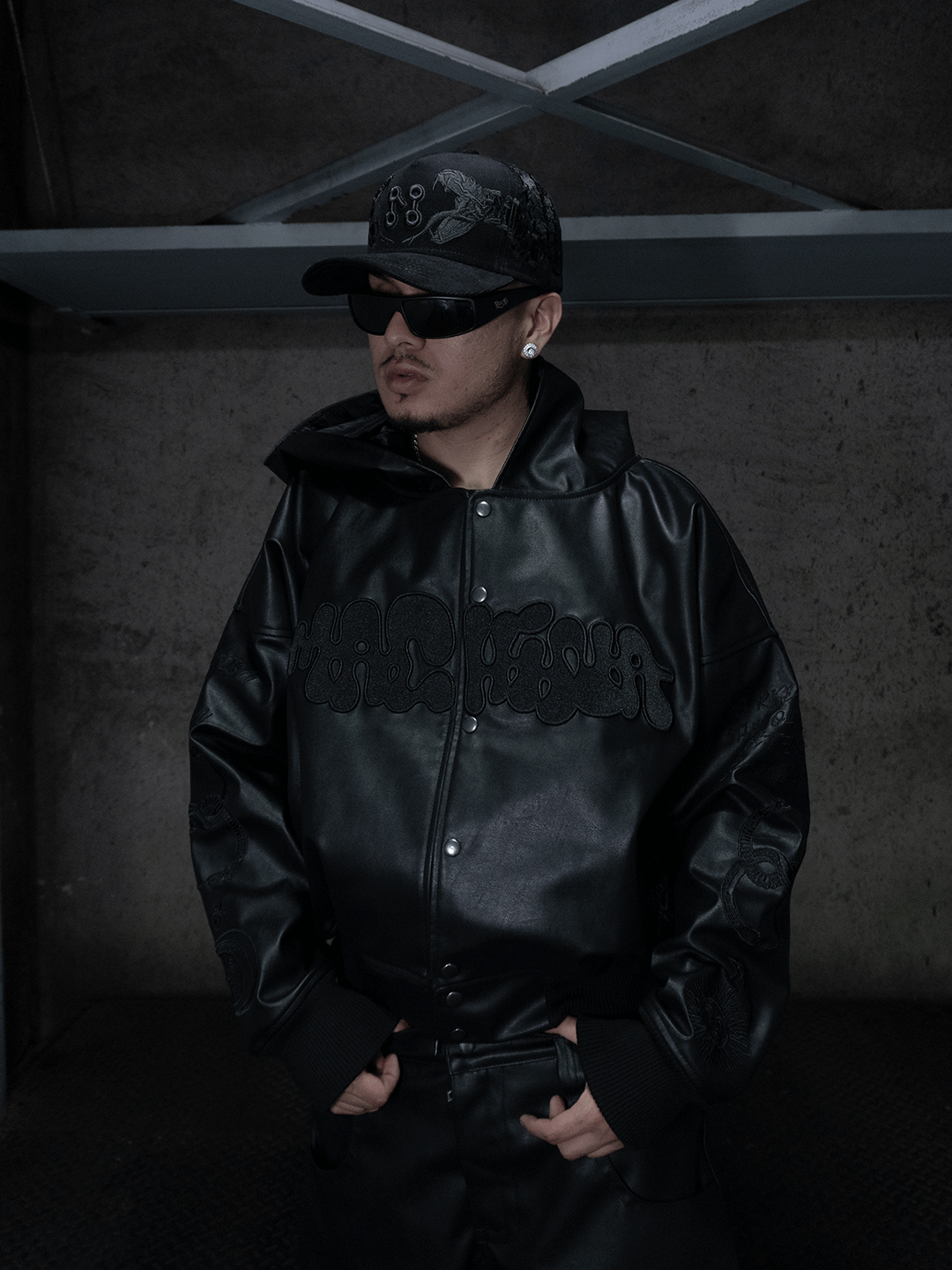 Wearing Machina - Wearable Technology's All-Black Eco Leather Bubble Set, a person stands indoors against a concrete wall in sunglasses and earrings, black cap on, hands clasped in front.