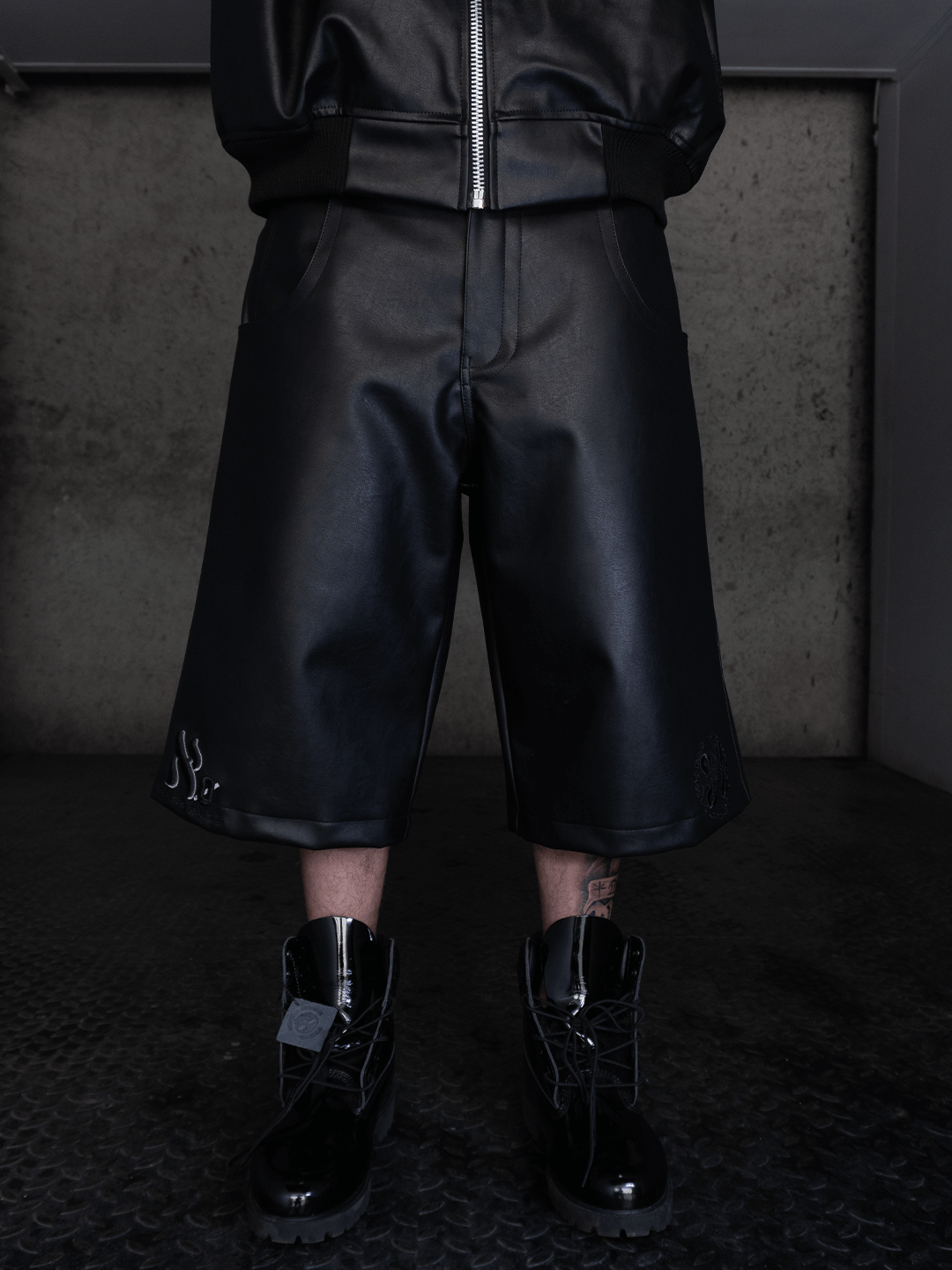 A person wears the All-Black Eco Leather Bubble Set by Machina - Wearable Technology, standing on a dark textured floor before a plain gray concrete wall. The set features knee-length shorts, boots, and a matching black jacket.