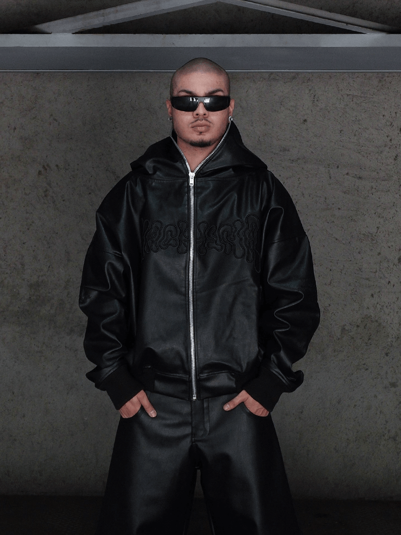 Dressed in the All-Black Eco Leather Osseous Full Zipper set by Machina - Wearable Technology, a person stands confidently against a concrete wall with hands in pockets, accessorized with black sunglasses and earrings.