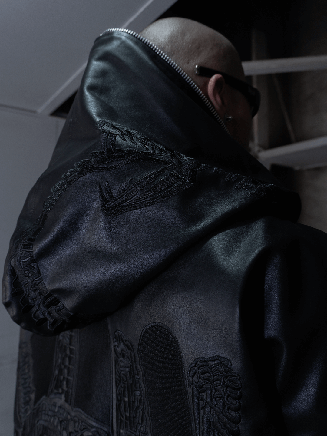 A bald person in sunglasses is seen from behind wearing Machina - Wearable Technology’s All-Black Eco Leather Osseous Full Zipper set, featuring embroidered designs and a large hood, standing in a dimly lit indoor space.