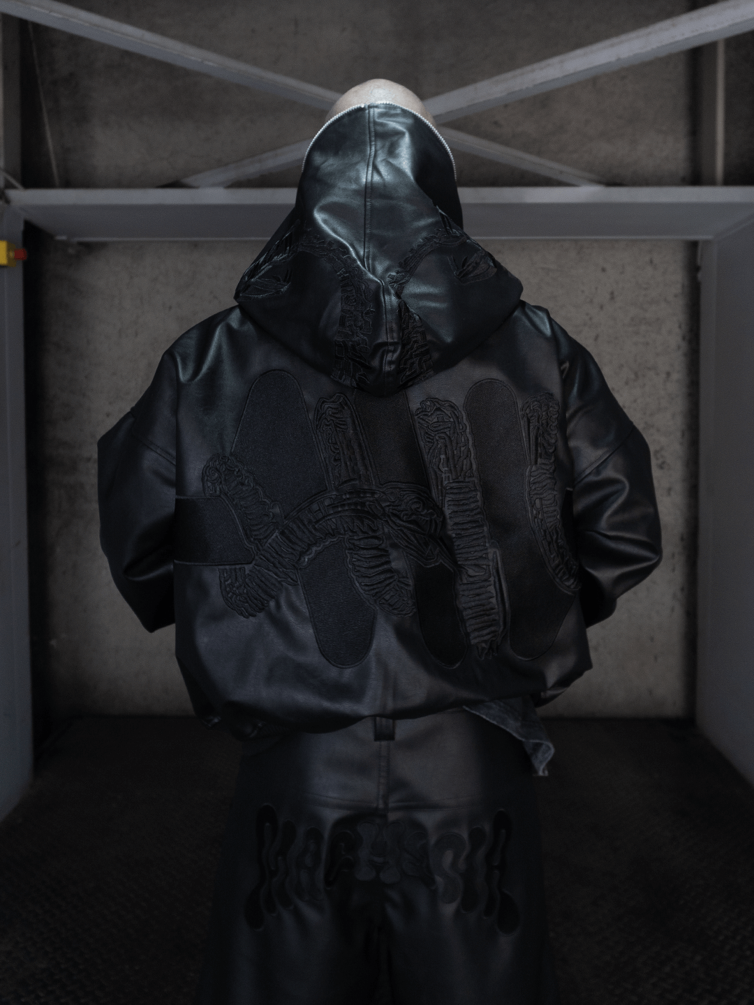 A person wearing the Machina - Wearable Technology All-Black Eco Leather Osseous Full Zipper set, featuring a hood and large textured handprint designs on the back, faces a concrete wall in an industrial space, evoking black ceremonial armor.