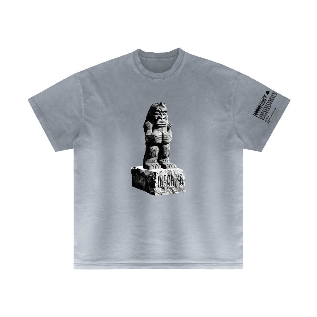 Introducing the "Testamentos de Piedra" by Machina - Wearable Technology: This gray T-shirt showcases an artistic image of a stone lion statue poised elegantly atop a carved pedestal, symbolizing an ancient alliance. The statue takes center stage on the shirt, complemented by text on the left sleeve. The design is sophisticated and monochromatic.