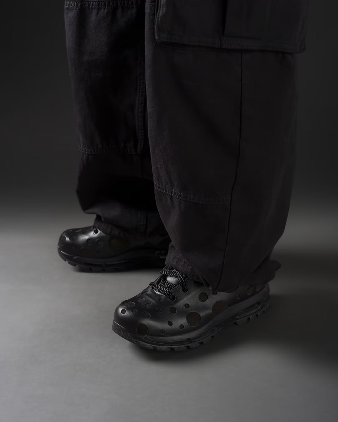 A person wearing wide, black pants and black sneakers with circular perforations stands on a gray surface against a dark background. Only the lower legs and feet are visible, highlighting the distinctive Air cushioning of the Machina Wearable Technology Air Max Goadome Low [Black].
