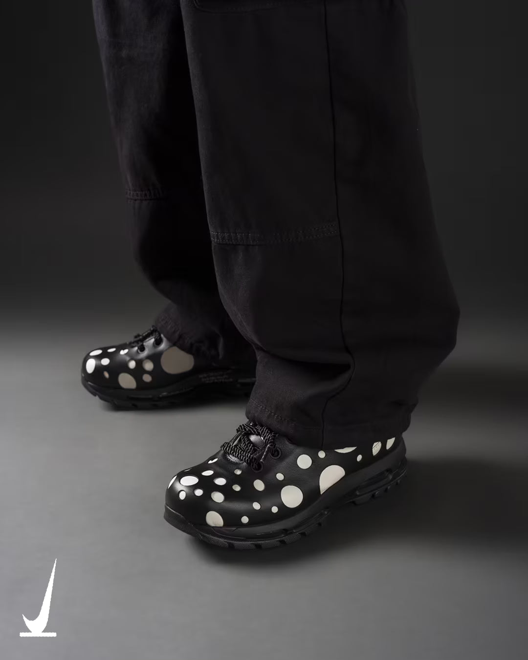 A person wearing black pants and Machina Wearable Technology Air Max Goadome Low shoes in Light Orewood Brown and Black, featuring large white polka dots, stands on a gray surface with a logo visible in the bottom left corner.