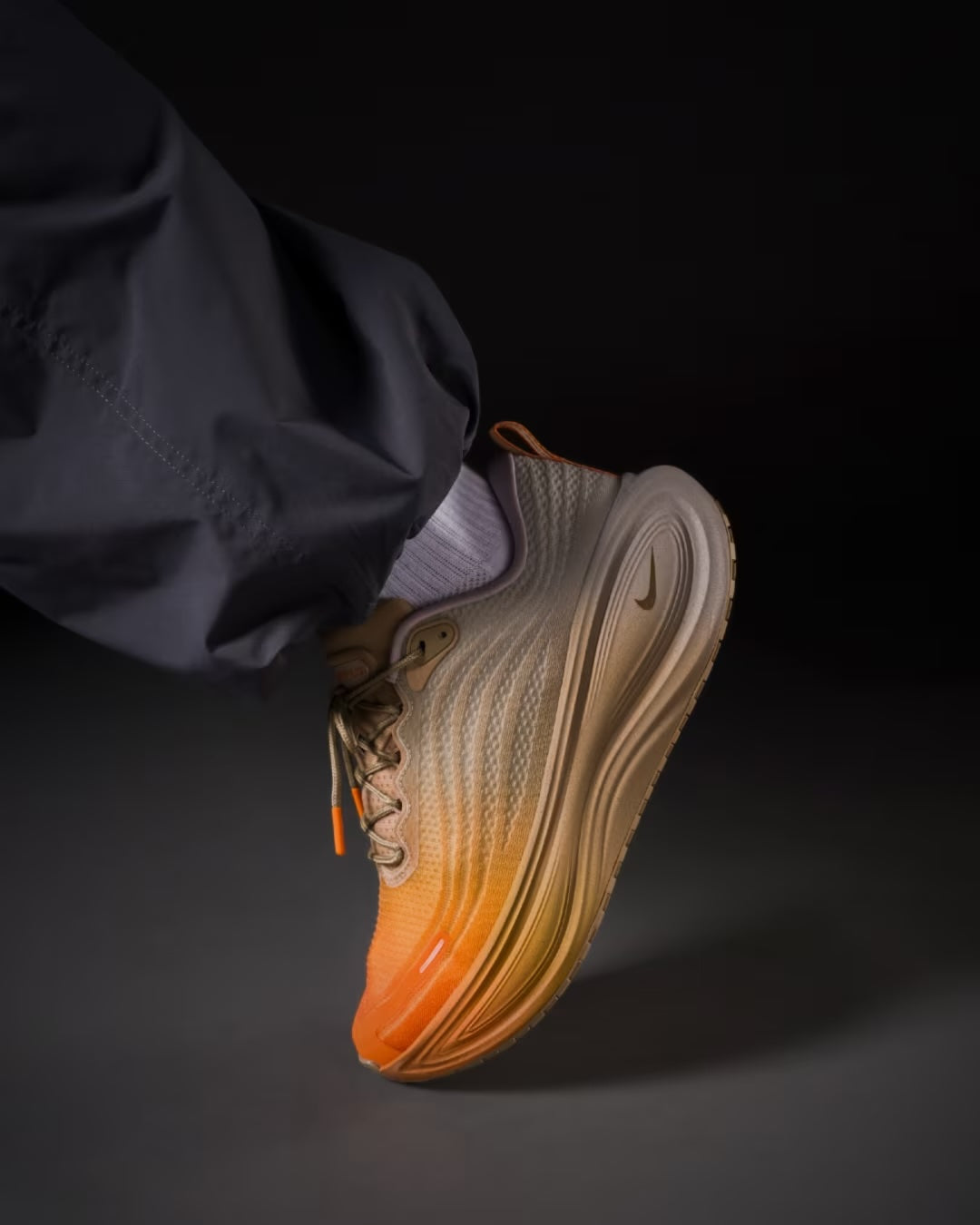 A person wearing black pants and white socks displays the Machina Wearable Technology Nike Vomero Plus “Cheat Day” running shoe, featuring an orange-to-brown gradient and a ZoomX midsole, set against a dark background.