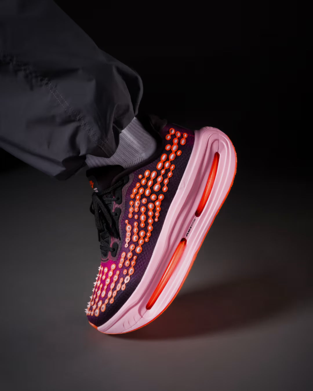 A close-up of The Nike Vomero Premium “Otaku Approved” sneaker from Machina Wearable Technology, featuring a black upper with pink and orange circular accents and a thick, light pink sole with a visible orange air cushion. Showcasing the ZoomX platform for maximum-cushion running, the foot is elevated while the wearer pairs dark pants with gray socks.