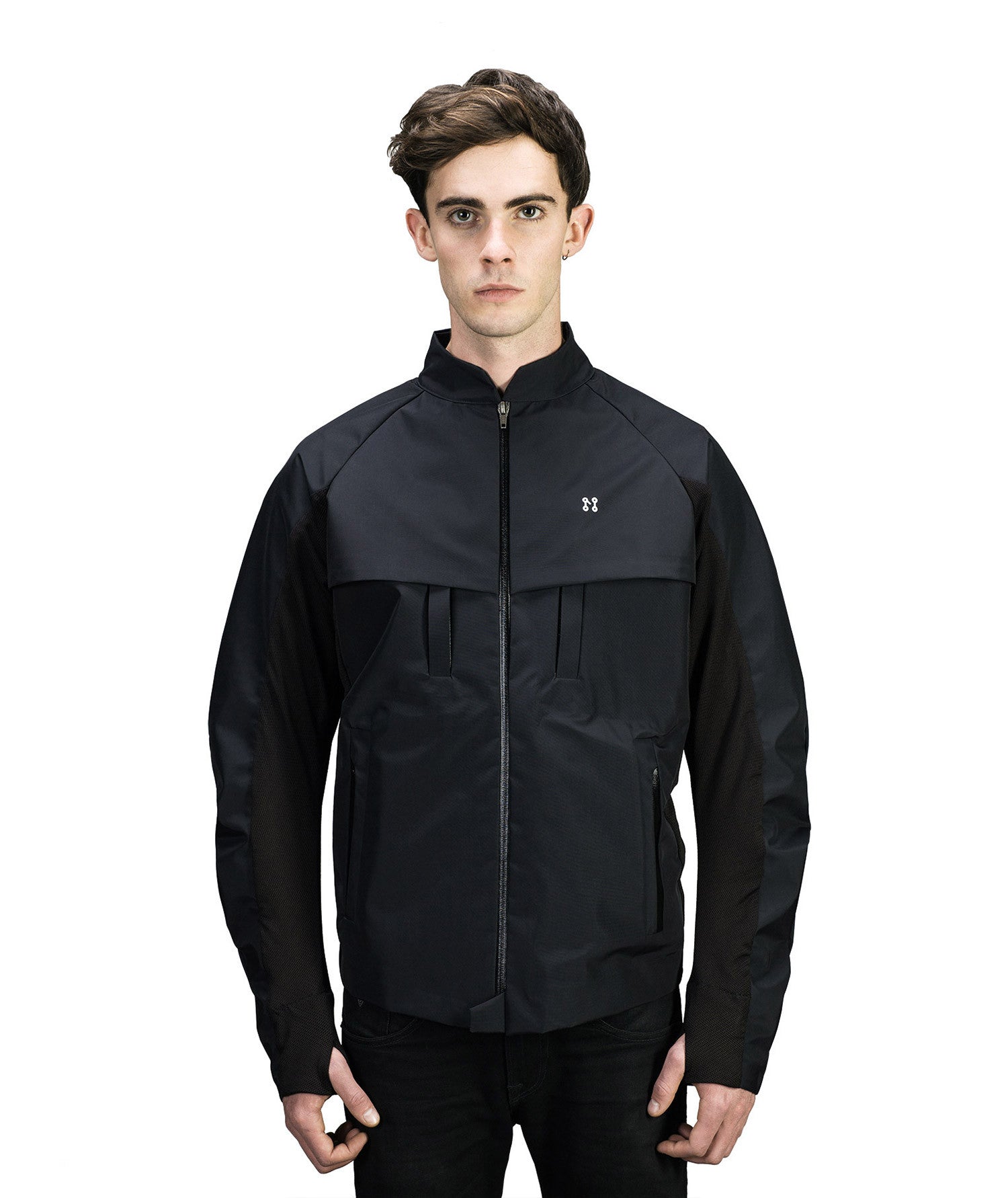 A young man with short brown hair is wearing Machina - Wearable Technology's MIDI Jacket, a black, high-collar piece subtly equipped with innovative body sensors. Against a plain white background and with his hands at his sides, the jacket displays a small logo on the chest and functions effortlessly as a MIDI controller to create music.