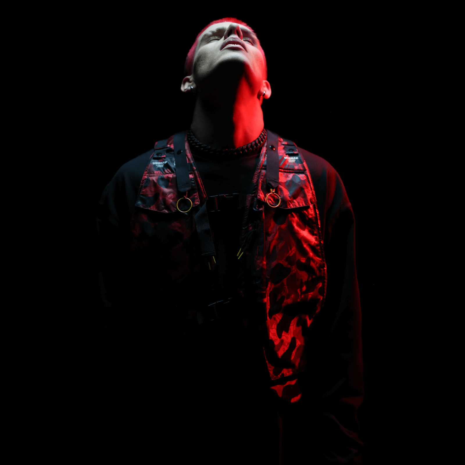 A person stands against a dark background, looking upward with eyes closed. They wear the Diablo Parachute Poncho by Machina - Wearable Technology, featuring an elegant black design with a red-patterned vest, complemented by an adjustable harness. Dramatic red and white lighting illuminates the scene, creating an atmosphere of intrigue and depth.