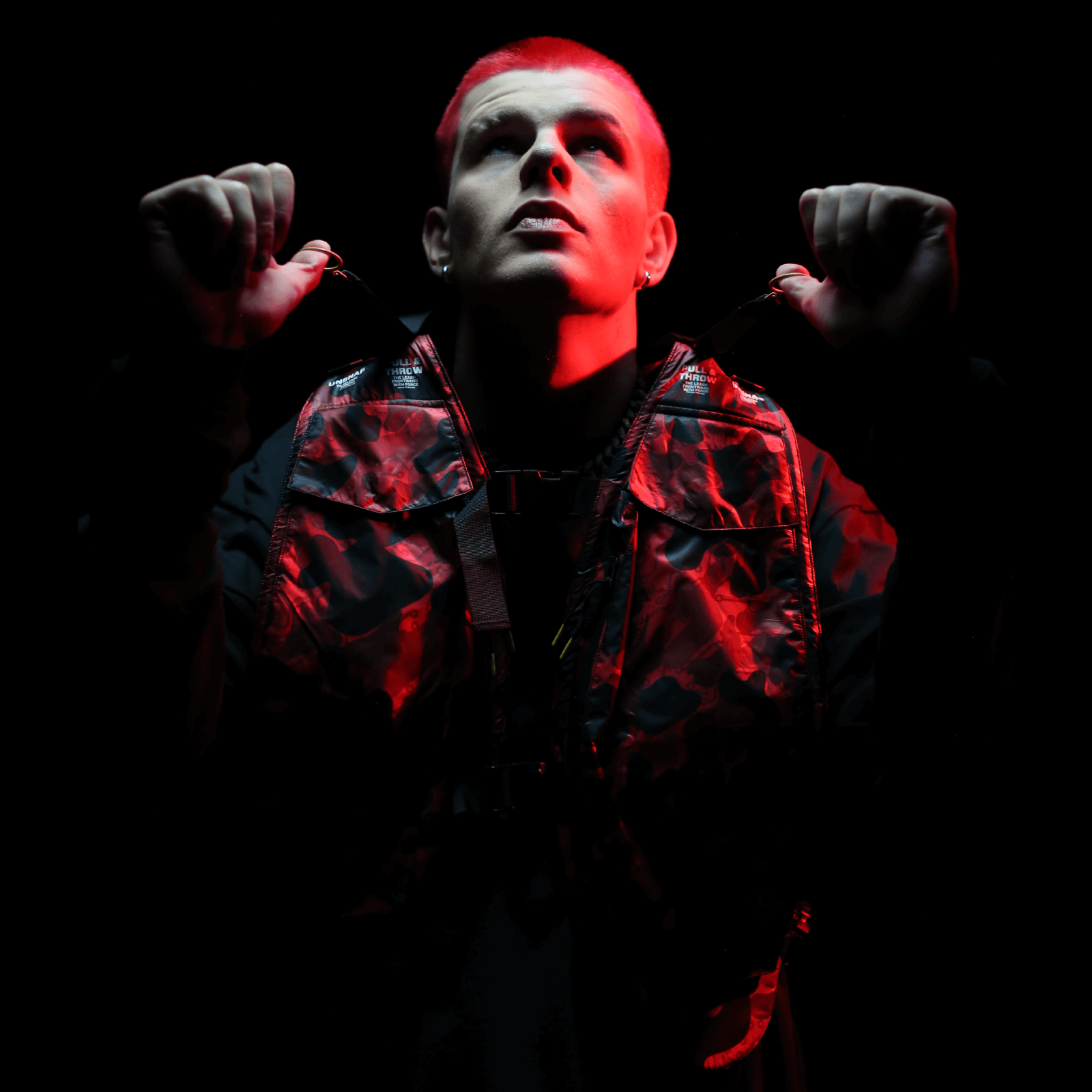 A person with short hair looks up, clad in a dark jacket and the sleek Diablo Parachute Poncho by Machina - Wearable Technology, which casts vibrant red reflections. The dramatic contrast of the illuminated figure against the dark background accentuates the poncho's distinctive design.
