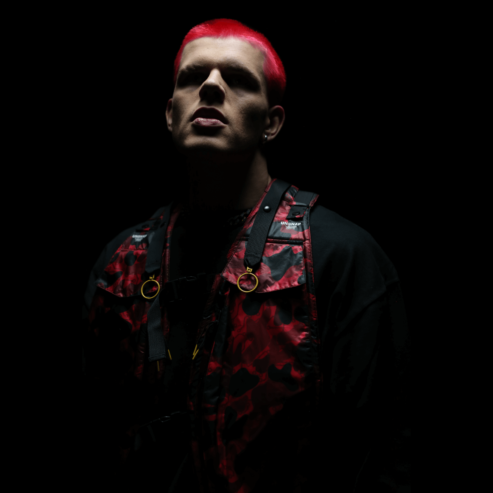 A person with bright red hair stands against a dark background, their ensemble completed by a Diablo Parachute Poncho from Machina - Wearable Technology over their black outfit and red-patterned vest. The lighting casts dramatic shadows across their face, enhancing the vibrant contrast of their attire.