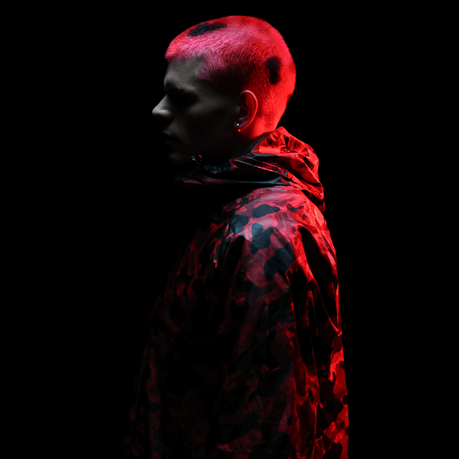 A person with short red hair and earrings is depicted in profile against a black background, wearing the Diablo Parachute Poncho by Machina - Wearable Technology. The poncho features a camouflage design reminiscent of an adjustable harness, highlighted by red light.