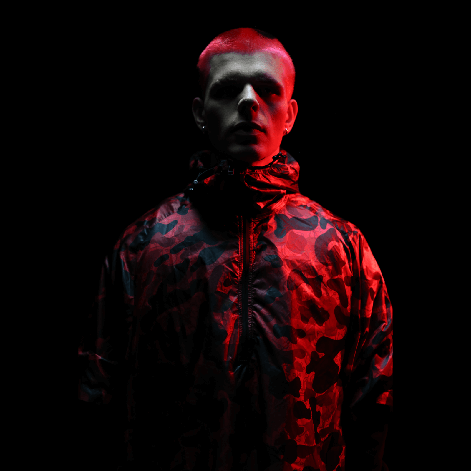 In a dimly lit setting, a figure is highlighted by red lighting while wearing a hooded jacket with a red and black pattern that resembles the Diablo Parachute Poncho from Machina - Wearable Technology. The lighting enhances the scene with a dramatic and intense atmosphere.