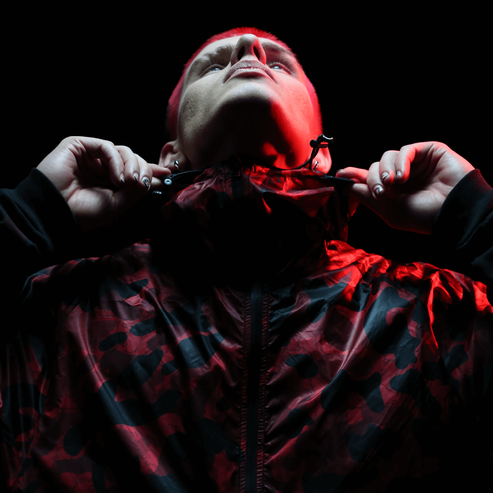 A person wearing the Diablo Parachute Poncho by Machina - Wearable Technology gazes upward against a dark background. Red lighting adds emphasis, casting striking shadows. The individual grips the collar of the poncho with both hands, highlighting its durable design.