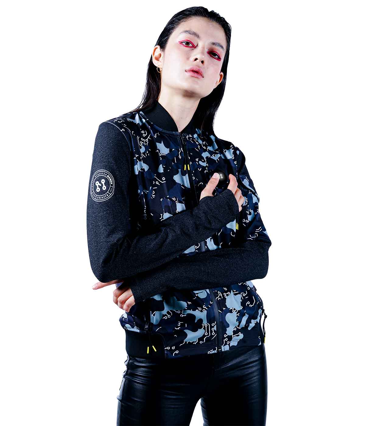 A person stands confidently with their arms crossed, wearing Machina - Wearable Technology's BLACK-CAMO BOMBER JACKET featuring a blue and black camo pattern with long dark sleeves. They pair it stylishly with black leather pants, slicked-back hair, and bold pink eye makeup. The scene captures the essence of fashion tech against a simple white background.