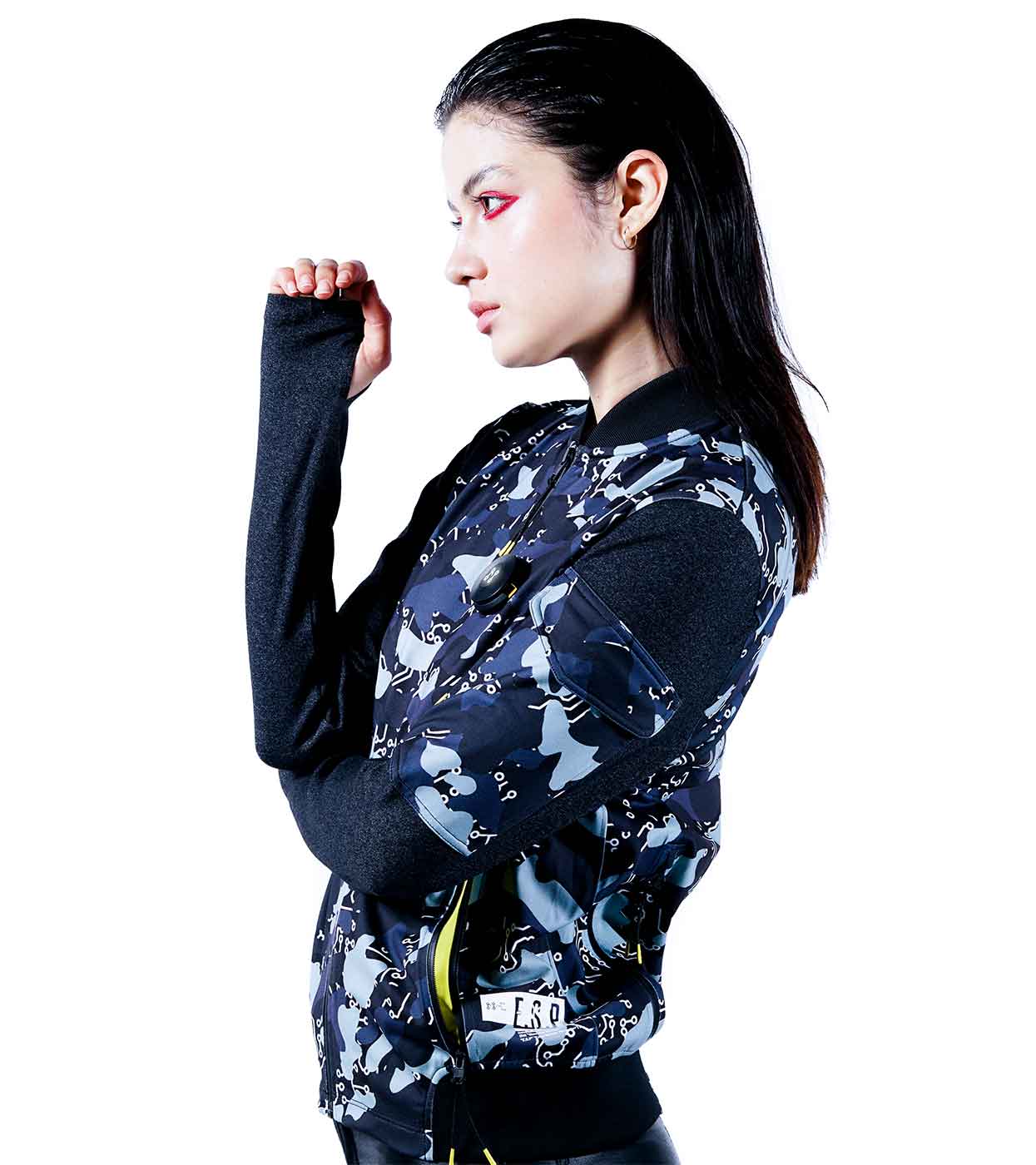 A person with long dark hair is wearing a BLACK-CAMO BOMBER JACKET by Machina - Wearable Technology, crafted from sustainable fabrics, over a black shirt. They pose sideways with their left arm slightly bent and hand near their face against a white background.