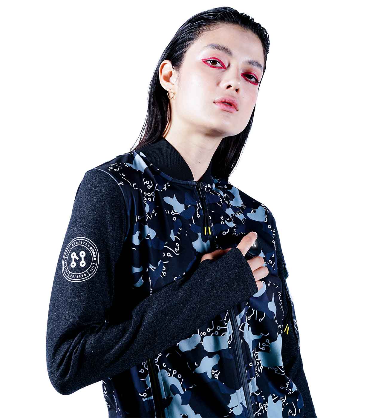 A person with long dark hair and bold pink eye makeup confidently gazes at the camera, wearing the BLACK-CAMO BOMBER JACKET by Machina - Wearable Technology. This women's smart bomber jacket is crafted from sustainable fabrics and showcases abstract designs in black, grey, and white, accented by a circular patch on the sleeve.