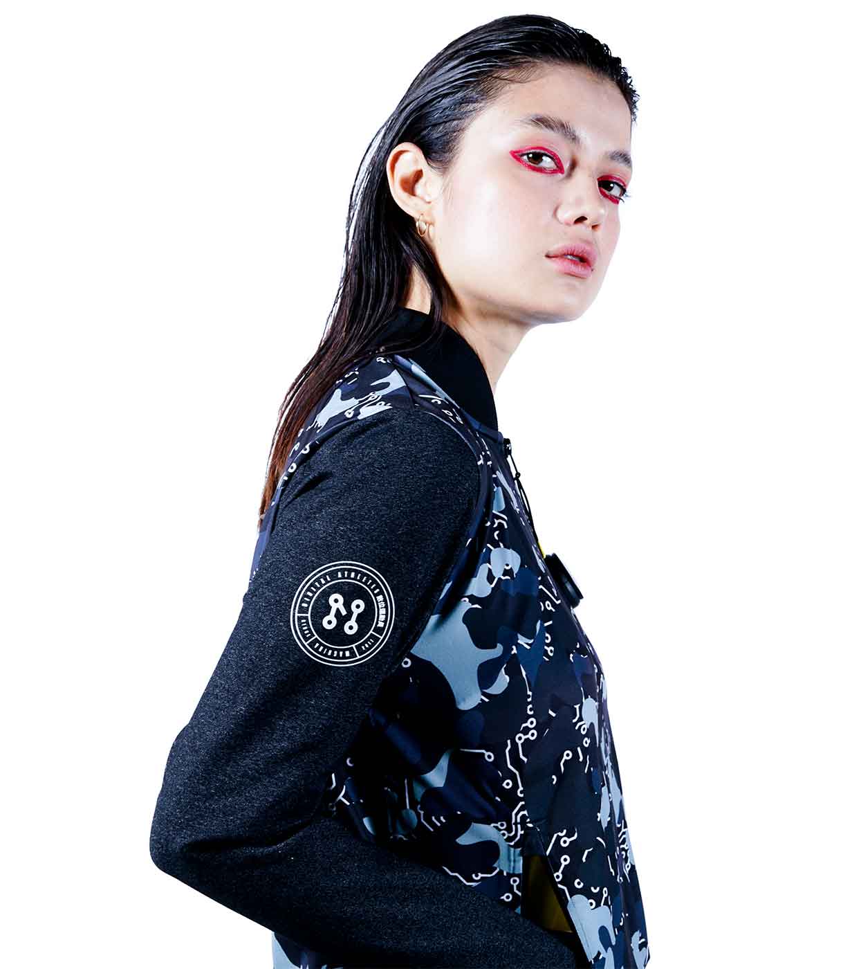 A model with stylish, wet-look hair and striking red eye makeup poses in the BLACK-CAMO BOMBER JACKET from Machina - Wearable Technology, highlighting its eco-friendly fabrics and black-and-gray camouflage design with a signature round emblem on the sleeve. The setting features a simple white background.