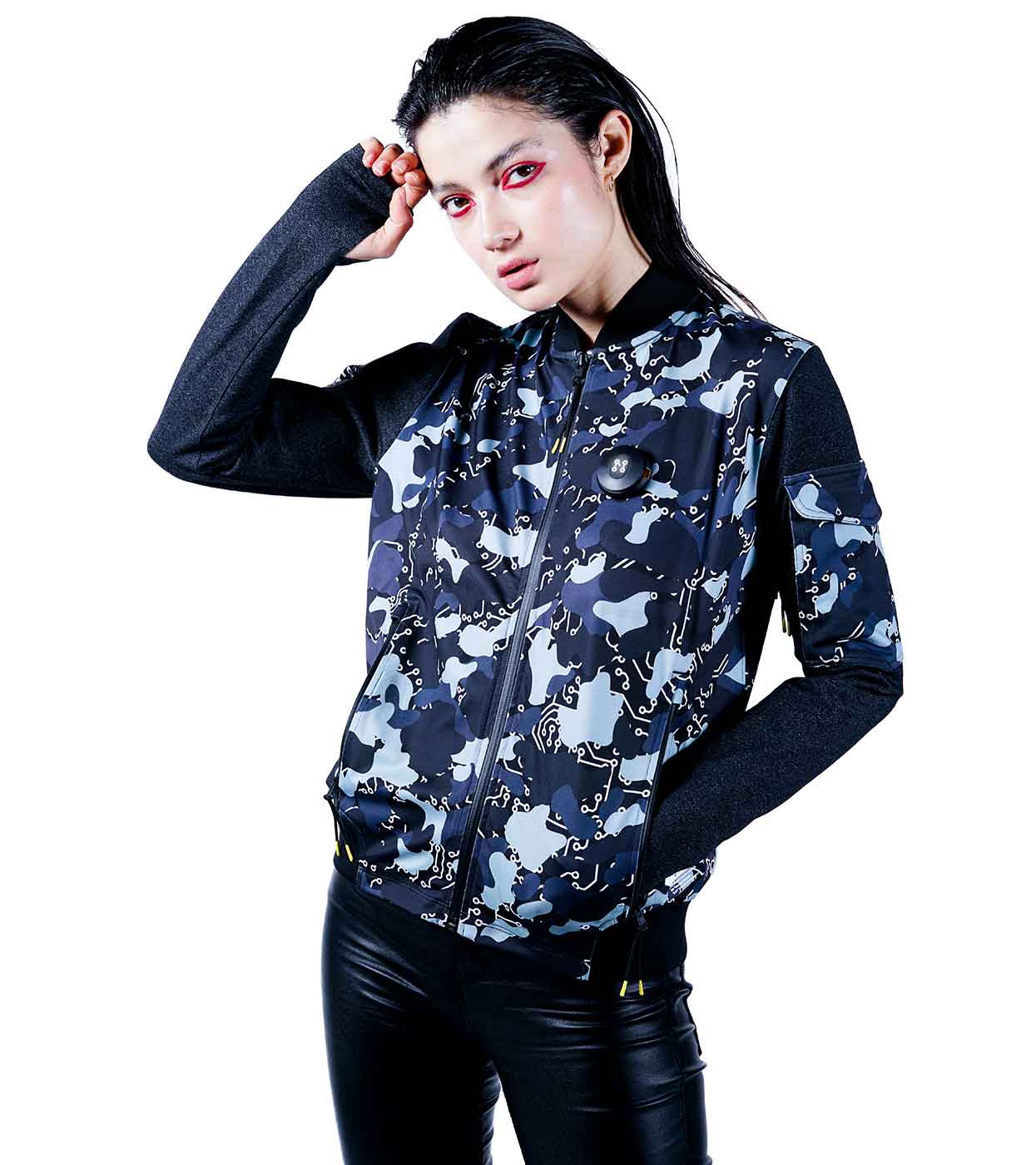 Dressed in Machina's BLACK-CAMO BOMBER JACKET, a person poses confidently with one hand on their hip and bright red eyeshadow. The jacket's blue and gray camo design with dark sleeves pairs seamlessly with their slicked-back hair and black leather pants.