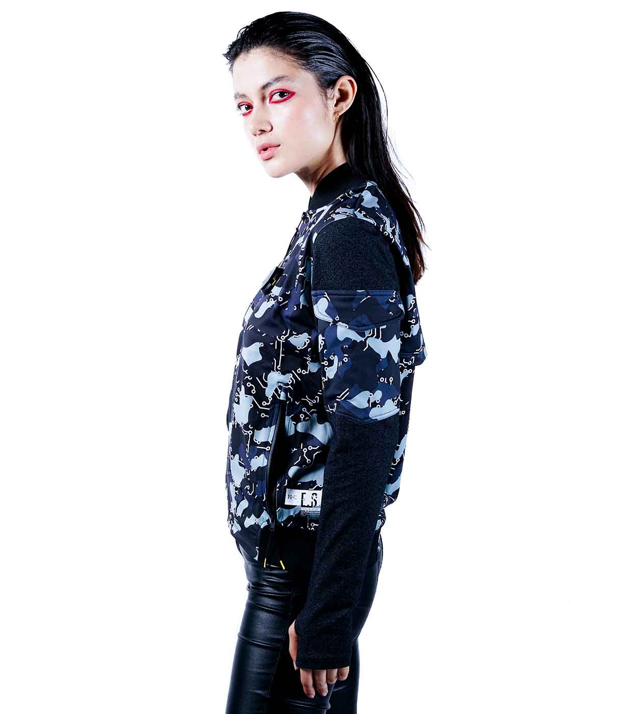A person with long hair and dramatic pink eye makeup is wearing the BLACK-CAMO BOMBER JACKET by Machina - Wearable Technology, artfully crafted from sustainable fabrics. The camouflage design pairs effortlessly with black pants as they stand against a plain white background, looking to the side.
