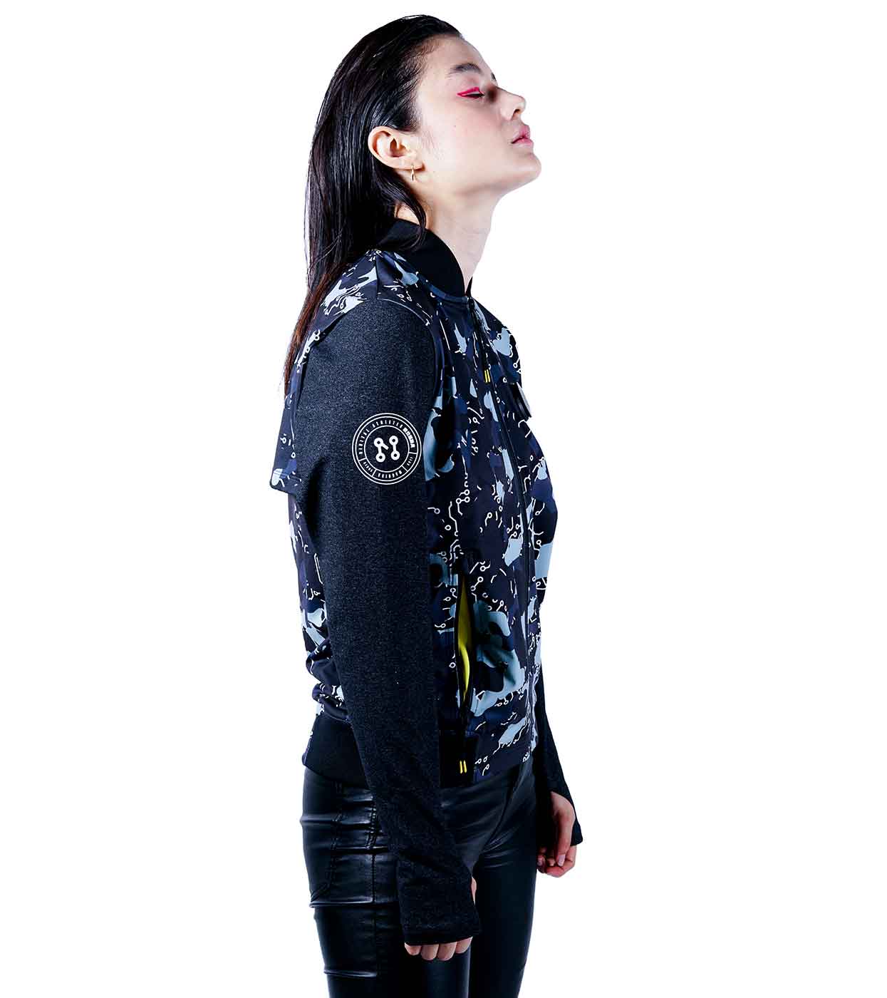 Against a white backdrop, an individual is captured in profile wearing Machina - Wearable Technology's BLACK-CAMO BOMBER JACKET, featuring a circular logo on the sleeve, paired with black leather pants. Their head tilts back gently, celebrating the fusion of fashion tech and sleek design.
