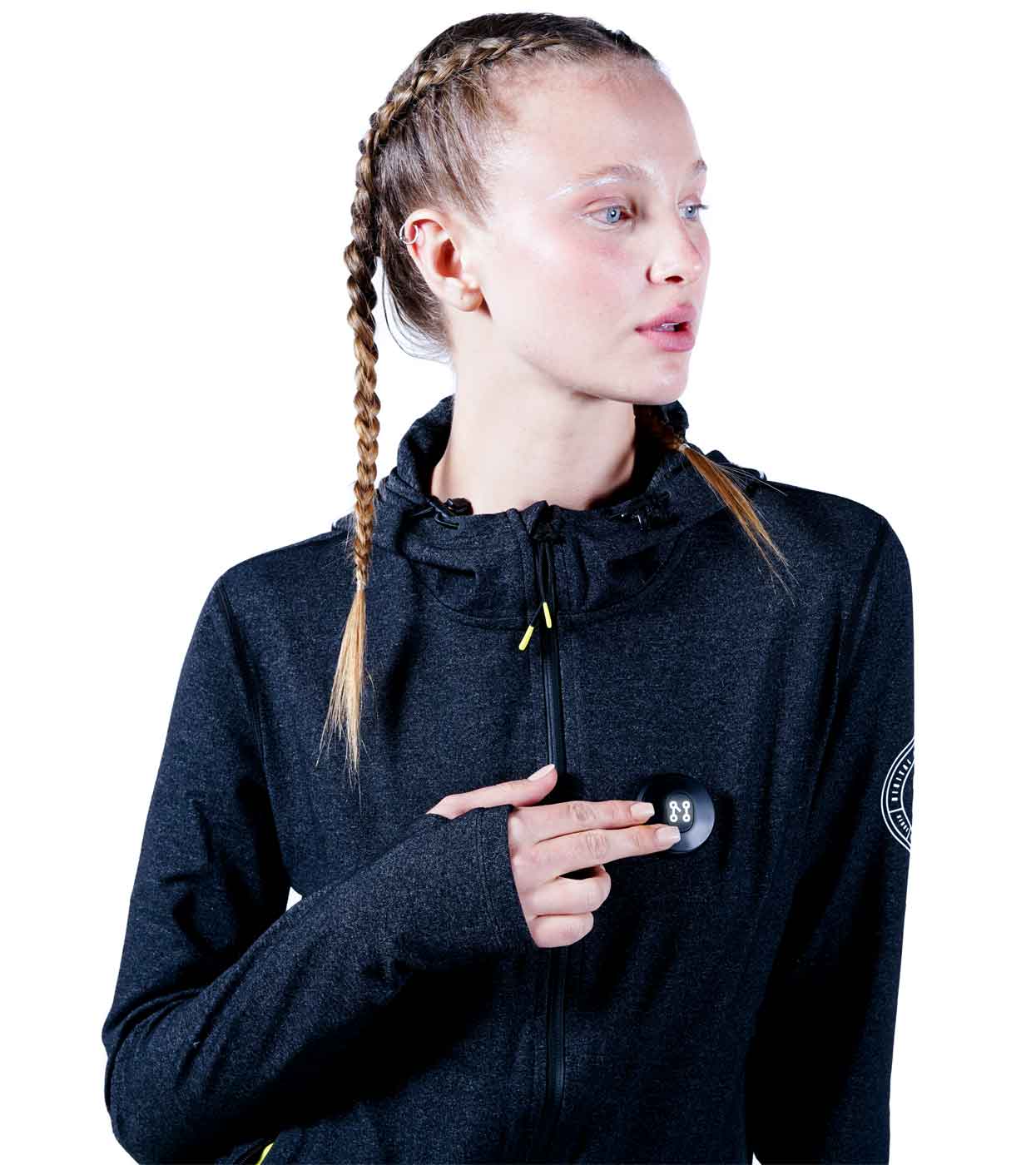 A person with braided hair, sporting a trendy MACHINA HOODIE from Machina - Wearable Technology, points to a small black device affixed to their chest. They gaze off to the side against a white backdrop.