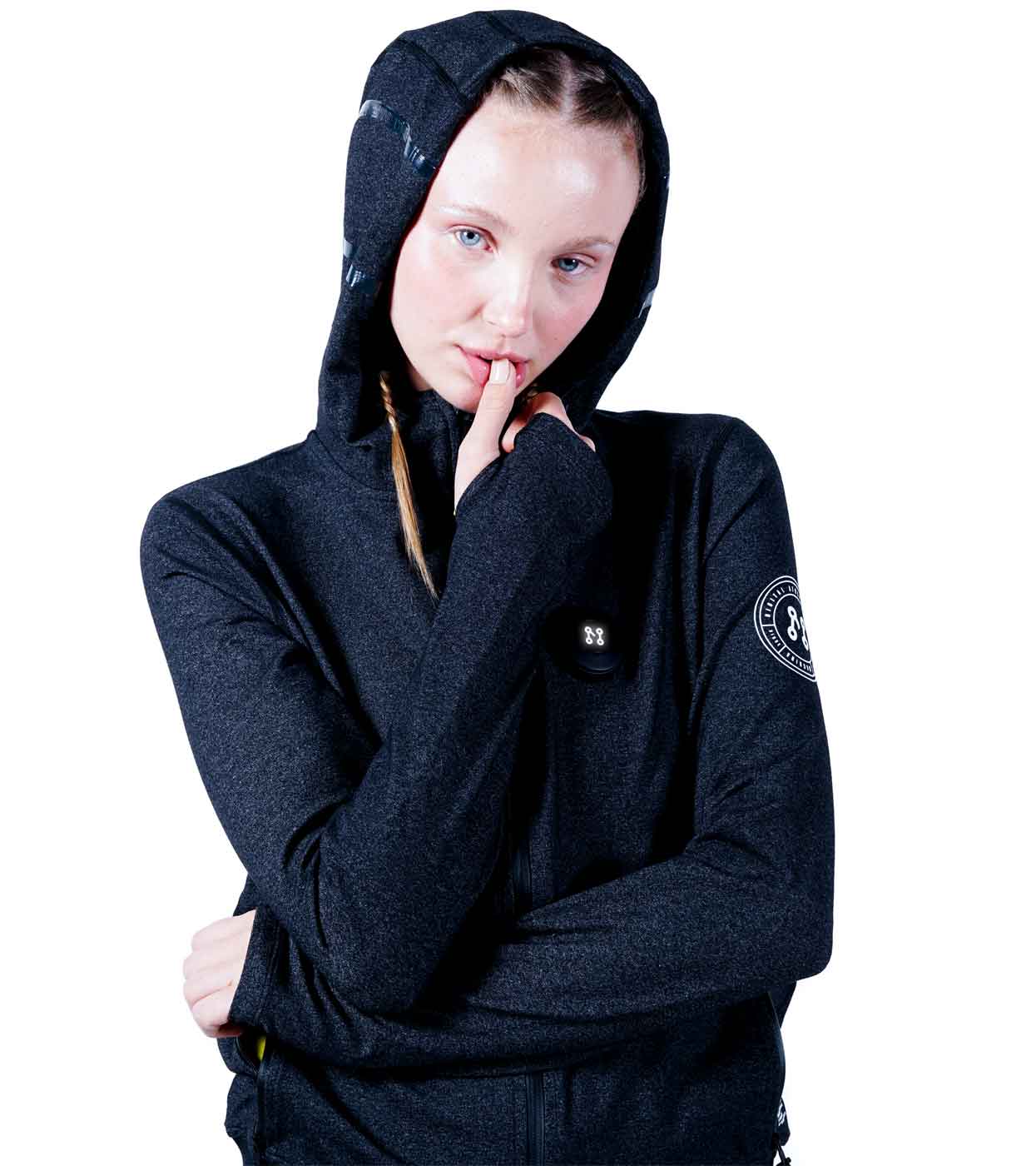 A person wearing the MACHINA HOODIE from Machina - Wearable Technology, featuring a distinctive patch on the sleeve, stands against a white background. Sporting two braids, they touch their lower lip with a finger, highlighting the perfect fusion of fashion tech and style.
