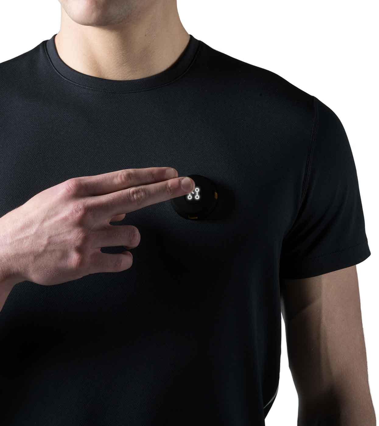 A person wearing a Machina Smart Black T-shirt, made from sustainable fabrics, is attaching a small, round electronic device with visible sensors or connectors to their chest. The person's face is not shown in the image.