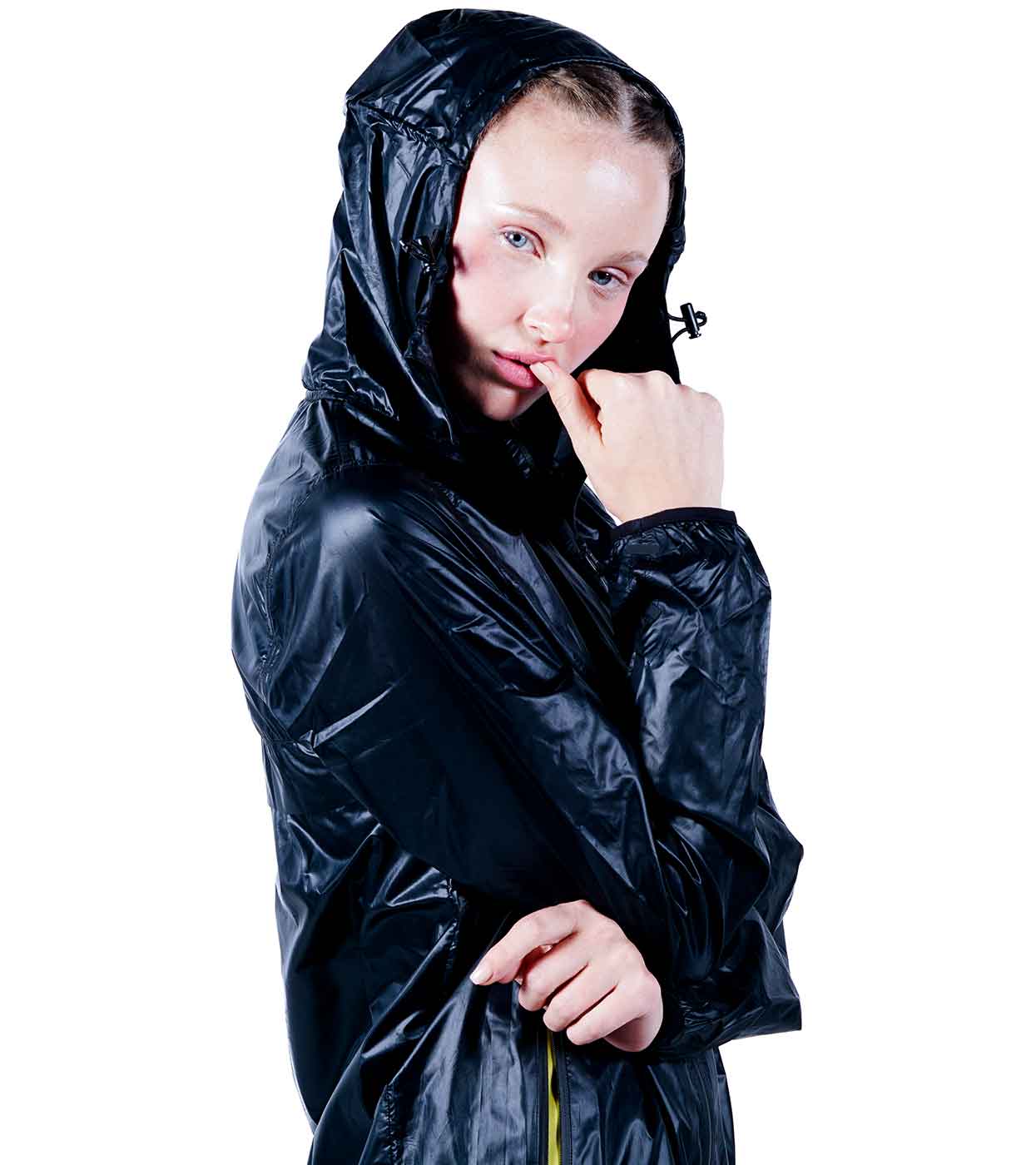 A person with light skin stands against a white background, wearing the BLACK WINDBREAKER by Machina - Wearable Technology. Their shiny black hooded jacket seamlessly combines fashion and functionality. One hand lightly touches their lips while the other arm is crossed.